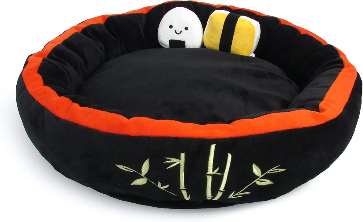 Small plush pet bed shaped like a black nori-wrapped sushi roll with a soft rice-white interior cushion, sized for cats or small dogs, shown with two matching plush sushi-piece crinkle toys arranged beside it